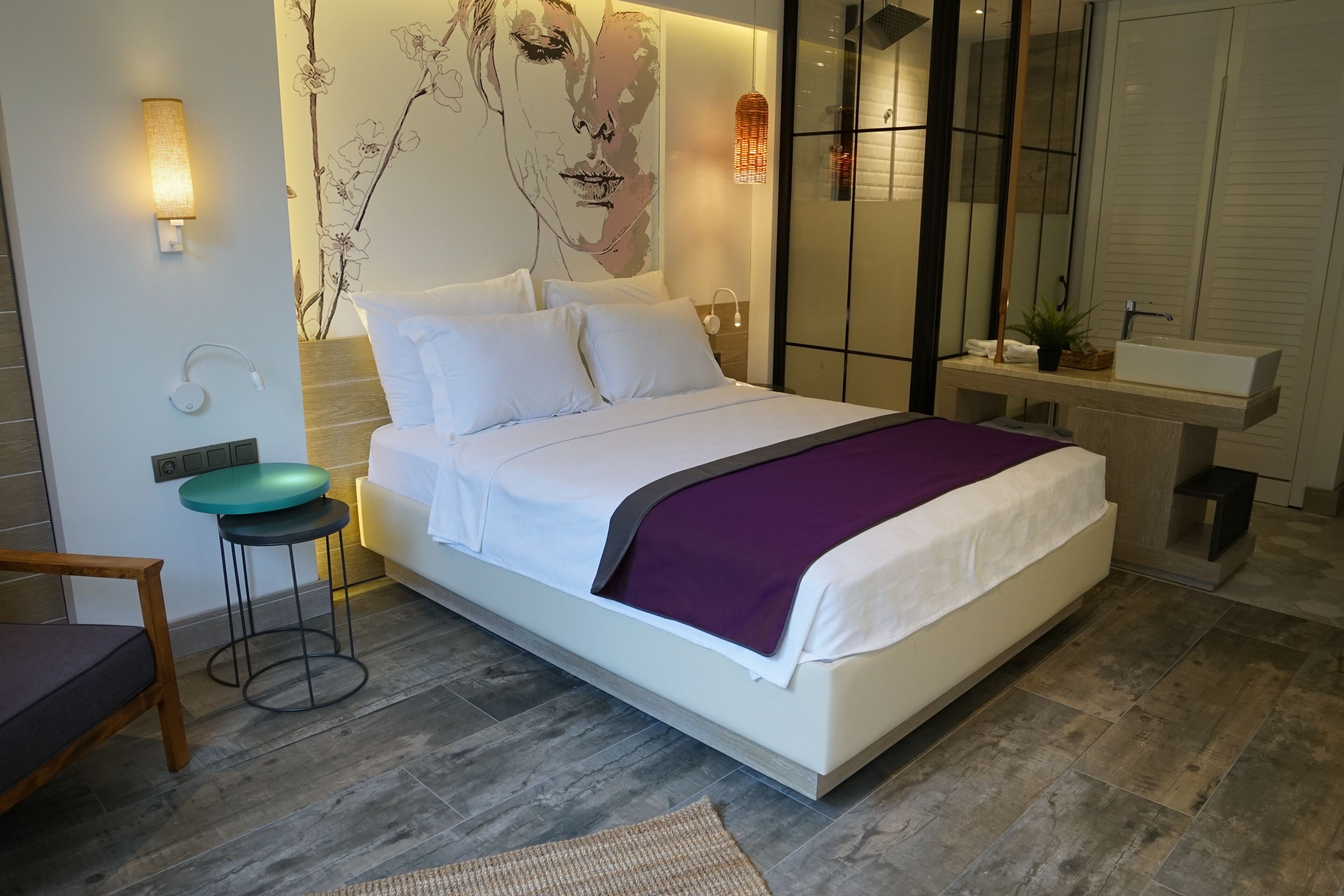 Ein modernes Schlafzimmer mit einem großen Bett, einem kleinen Hocker, einem Stuhl, einem Nachttisch, einer Wandlampe und einem Badezimmer mit Waschbecken und Spiegel.