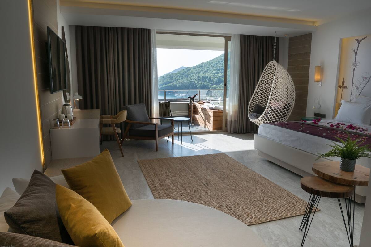 Una habitación de hotel moderna con una cama grande, un área de descanso con una silla y una mesa, un balcón con vista al mar y un televisor montado.