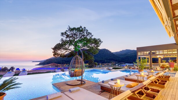 An infinity pool with lounge chairs and a hanging chair, surrounded by a wooden deck and a view of the sea and mountains.