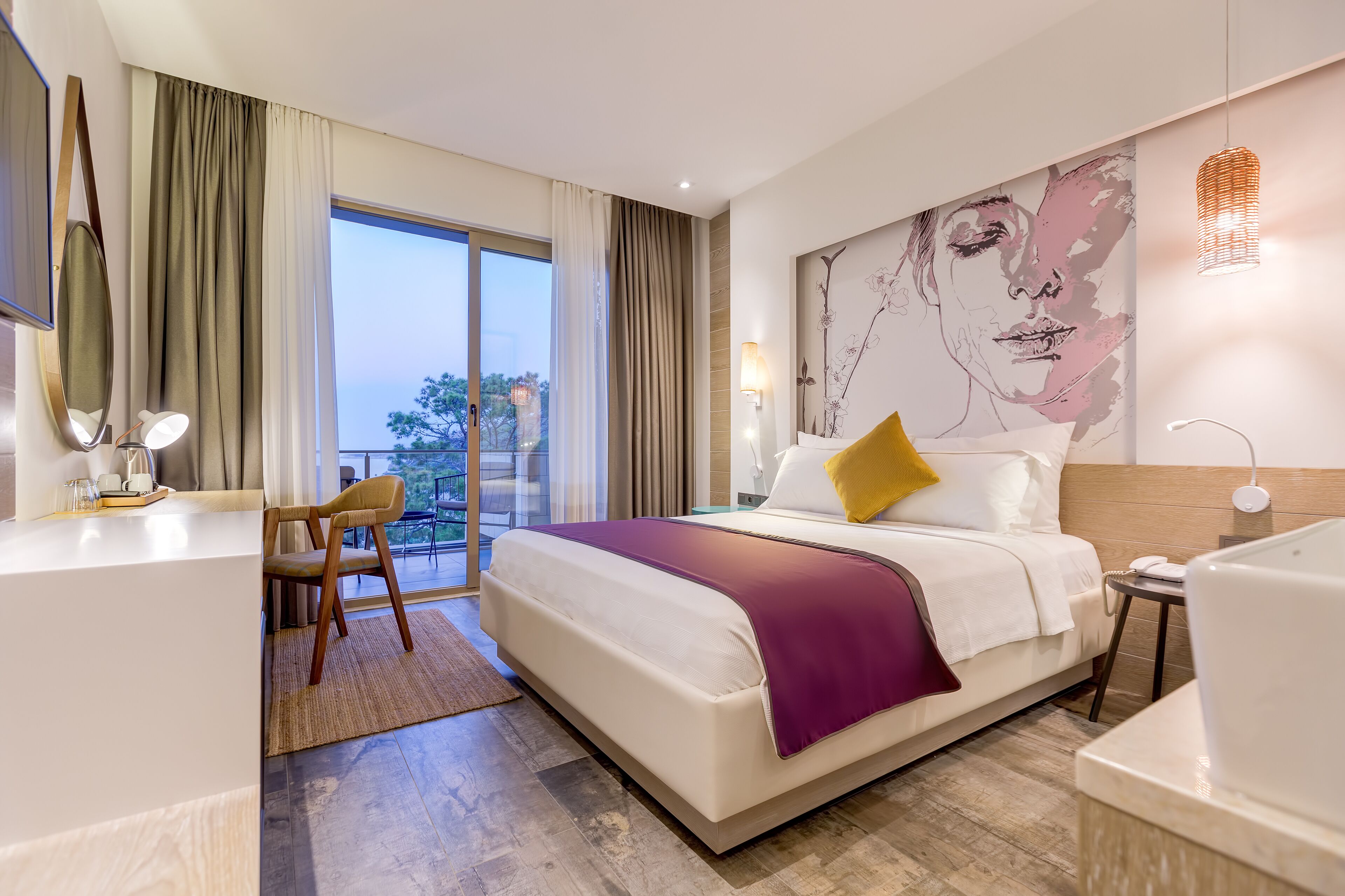 A modern hotel room with a large bed, a desk, a chair, a balcony with a view, and a large artwork on the wall.