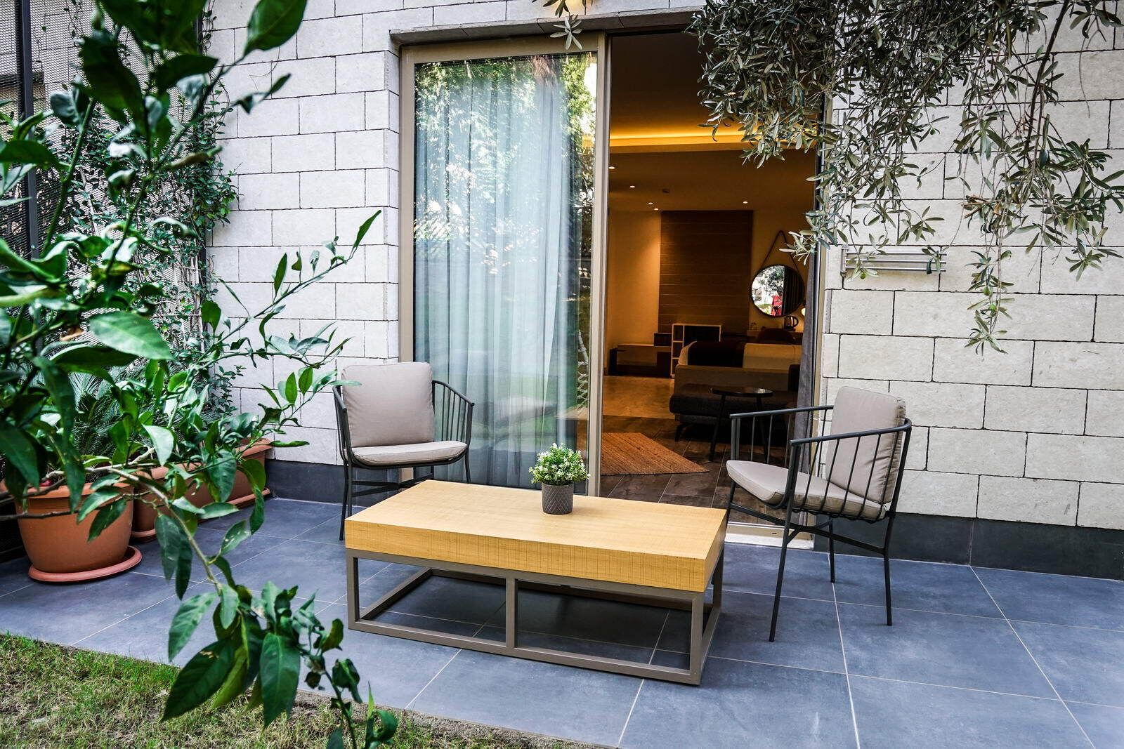 A modern outdoor seating area with a wooden table and two chairs, surrounded by potted plants and a glass door leading to an interior space.