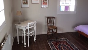 Een kamer met een houten vloer, een wit bureau met een lamp, een houten stoel, een bed met een paars kussen, en drie ingelijste foto's aan de muur.