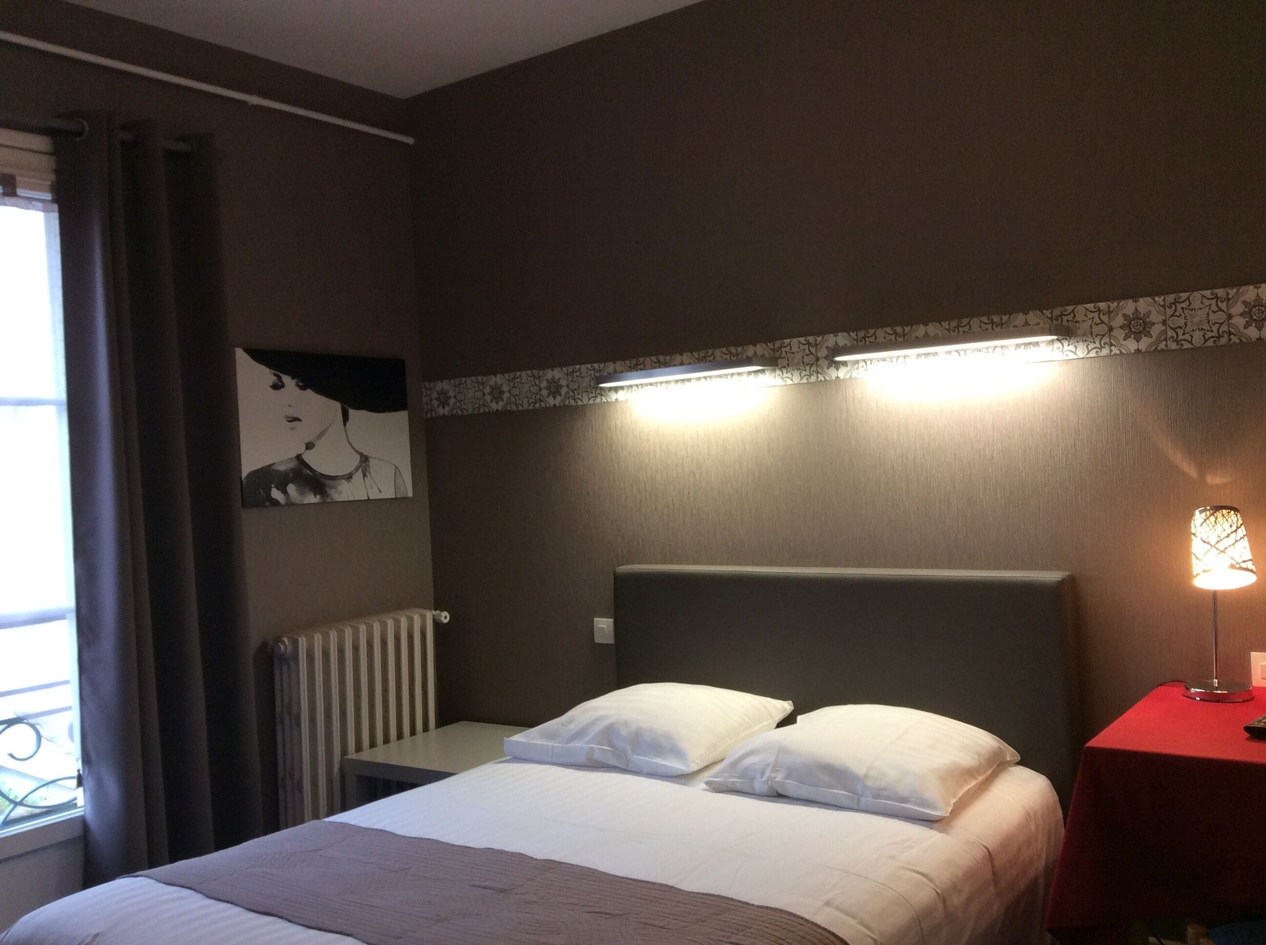 A hotel room with a bed, a nightstand, a lamp, a radiator, and a window with curtains.