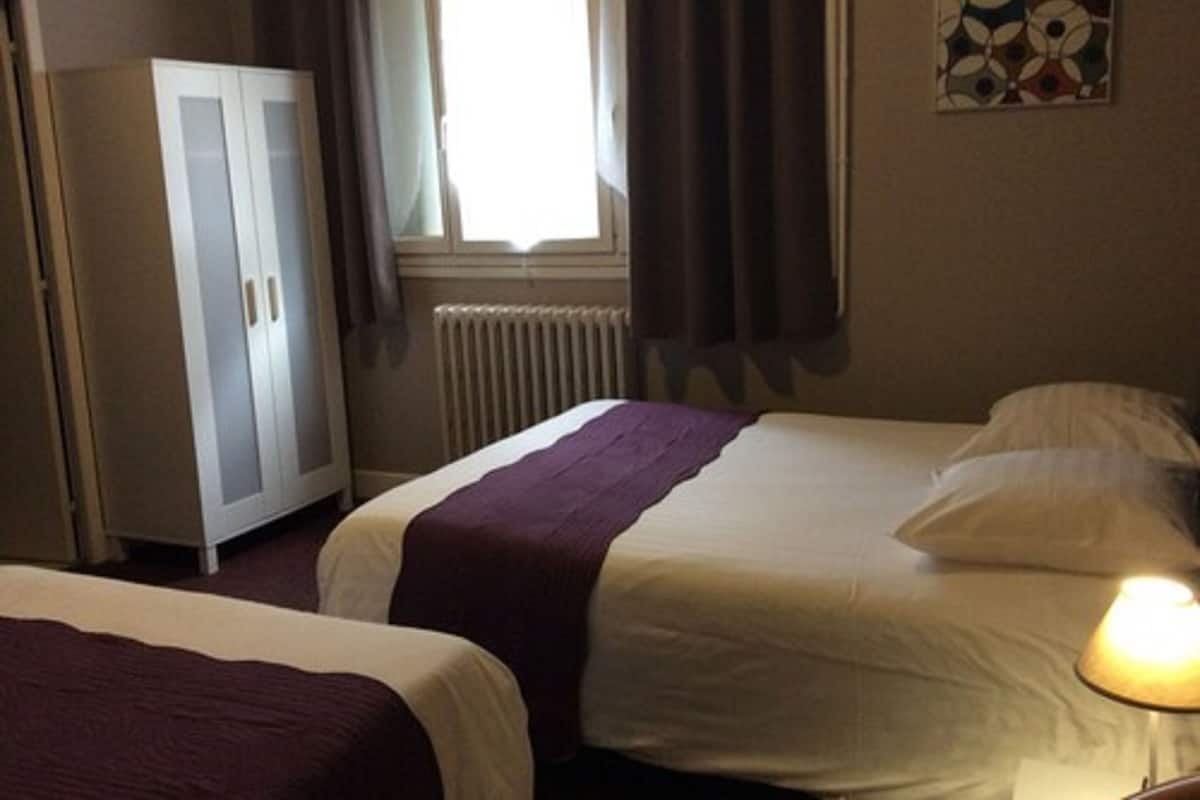 A hotel room with two beds, a wardrobe, a radiator, a window with curtains, and a wall-mounted artwork.