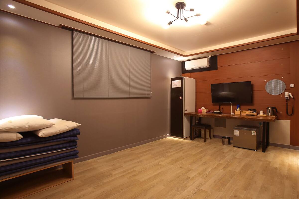 A hotel room with a wooden desk, a television, a small refrigerator, and a stack of pillows.