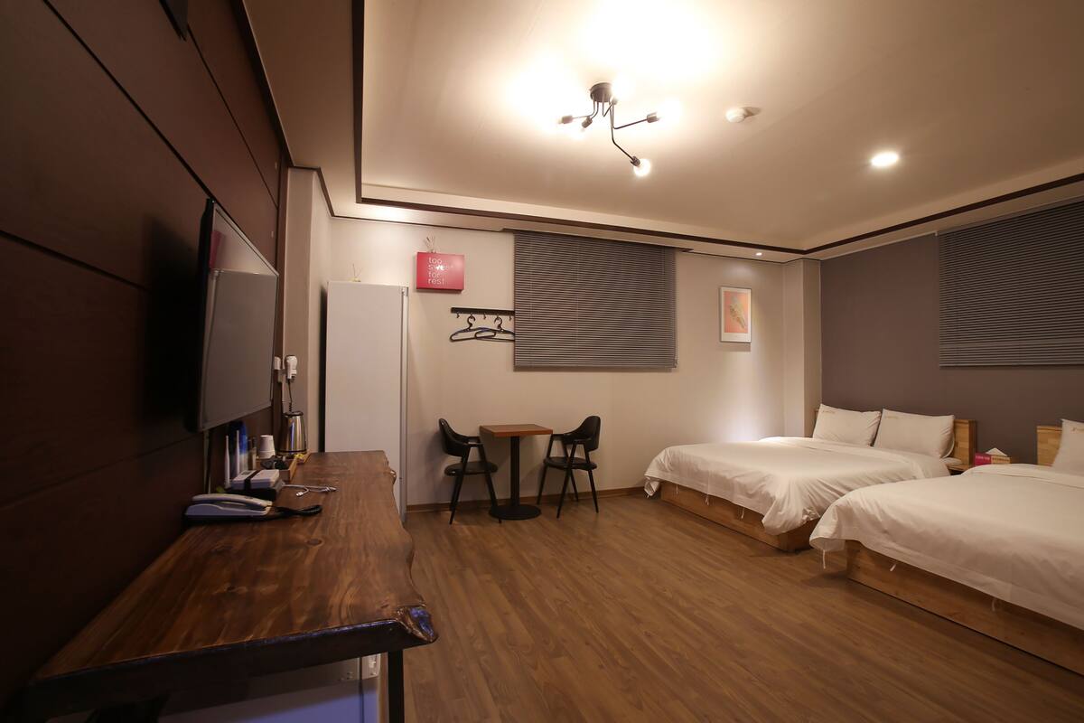 A hotel room with two beds, a wooden desk, a small table with chairs, a TV, and a ceiling light fixture.
