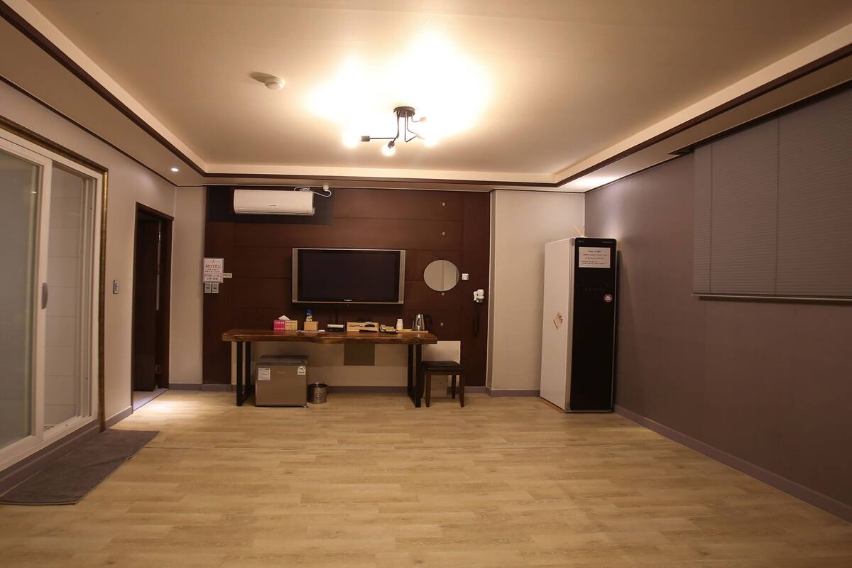A hotel room with a wooden floor, a flat-screen TV mounted on the wall, a desk with a chair, a refrigerator, and a door leading to another room.