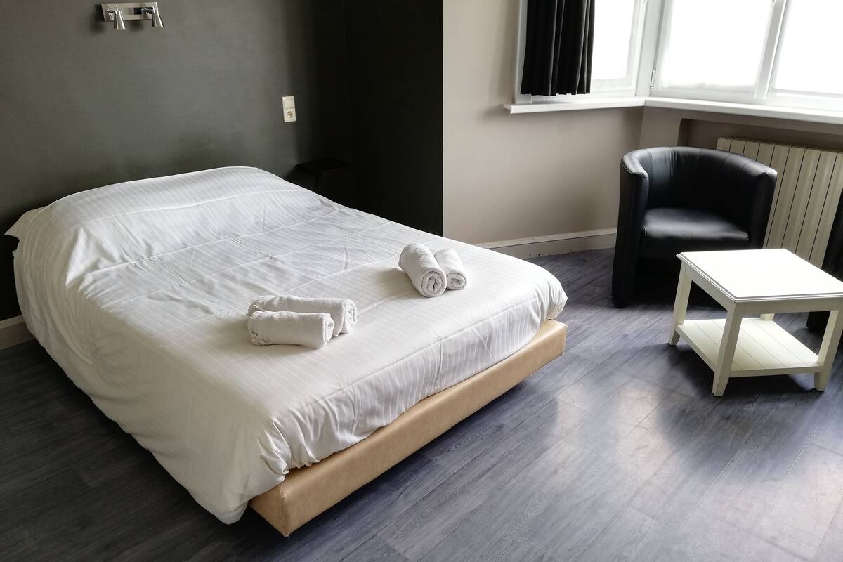 A neatly made bed with white linens and towels, a small white table, a black chair, and a window with blinds.