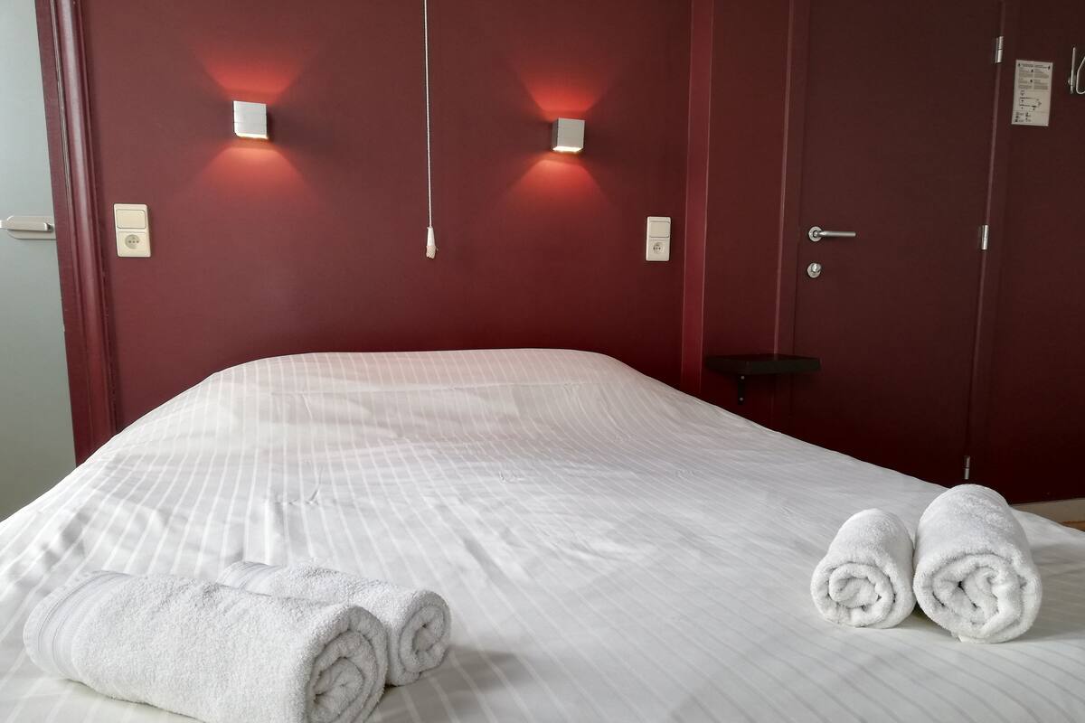A neatly made bed with white linens and two rolled towels, a red wall with two wall-mounted lights, and a door with a handle.