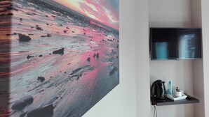 Una habitación con un cuadro abstracto grande en la pared, un televisor de pantalla plana montado en la pared y una pequeña estantería con una cafetera, una botella de agua y una taza.