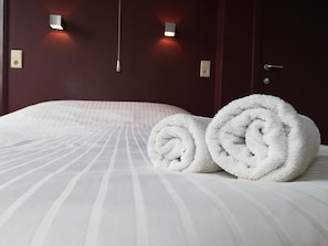Una cama bien tendida con toallas blancas enrolladas, un cabecero de madera oscura y dos lámparas fijadas a la pared.