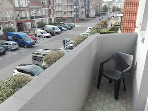 Un balcón con una silla, con vista a una calle donde se ven coches aparcados y una furgoneta.