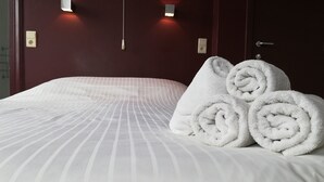 Una cama bien hecha con sábanas blancas y toallas enrolladas, un cabecero de madera oscura y dos lámparas fijadas a la pared.