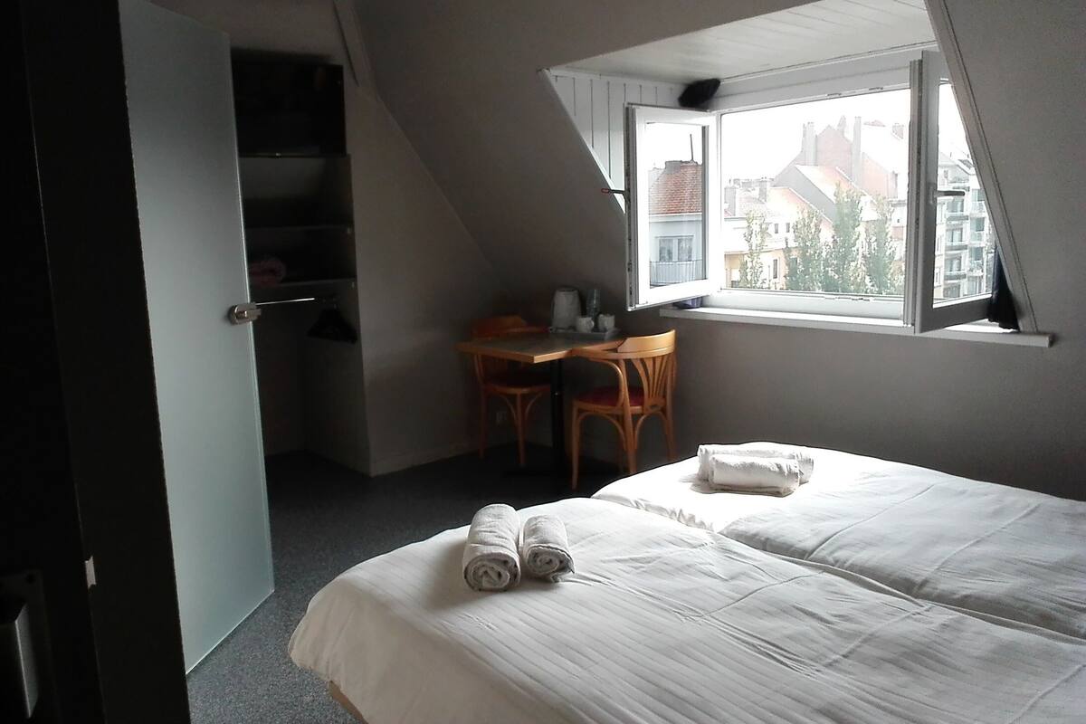 A bedroom with a bed, a desk, and a chair. There is a window with a view of buildings outside.
