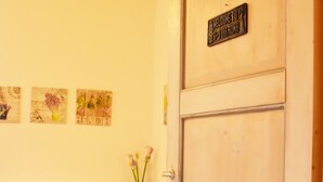 Una habitación con puerta de madera, un sofá blanco con cojines amarillos, una mesita con un bol y un florero con flores.