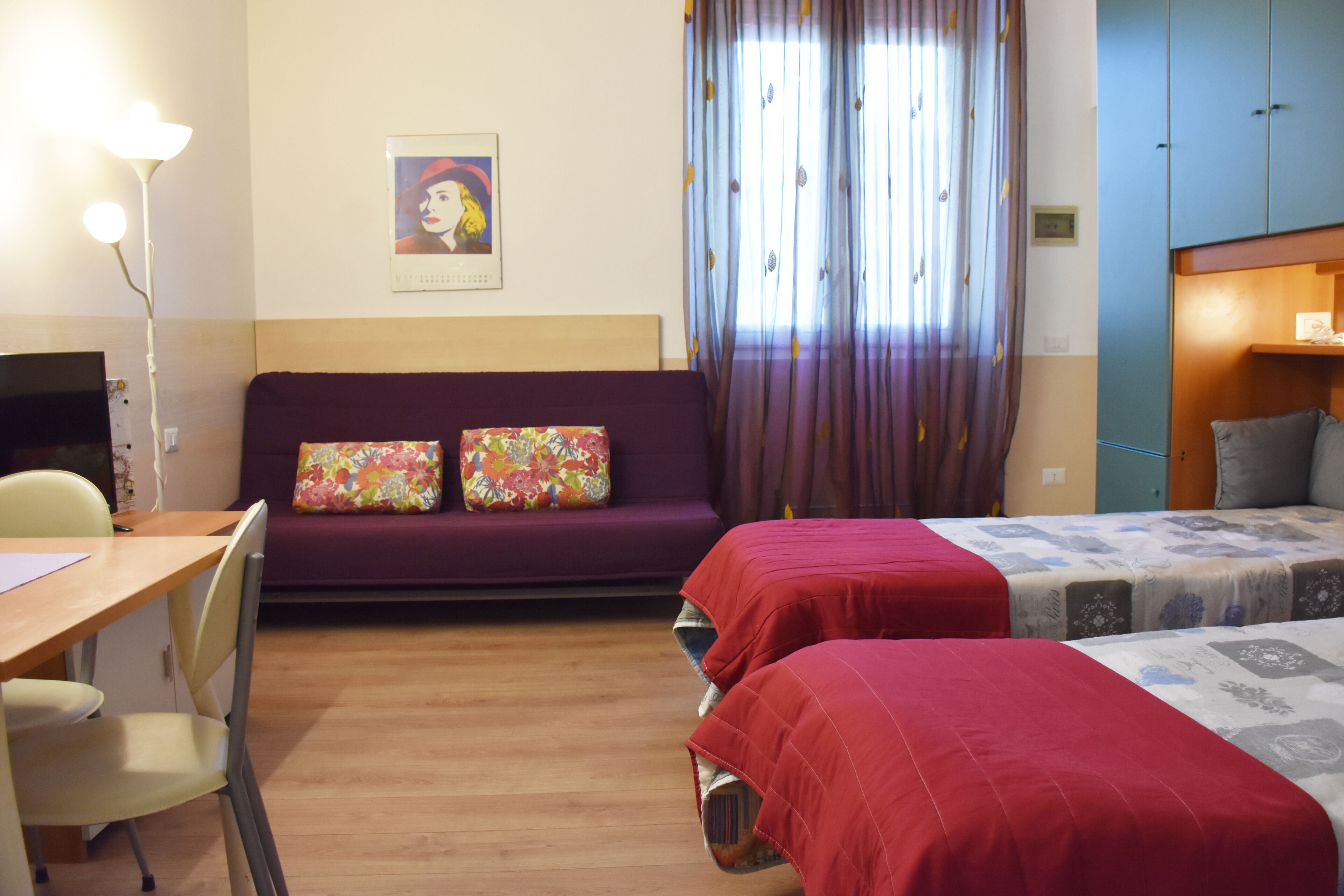 A hotel room with a wooden floor, a sofa with floral cushions, two single beds with red and patterned bedspreads, a desk with a chair, and a television.