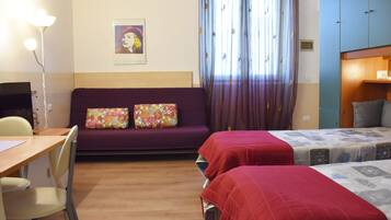 A hotel room with a wooden floor, a sofa with floral cushions, two single beds with red and patterned bedspreads, a desk with a chair, and a television.