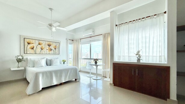 A modern bedroom with a bed, a ceiling fan, a large window with curtains, and a view of the city. - Charming • Lovely view • Beach • Pool 1304 (Cartagena)
