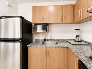 Una cocina con alacenas de madera, un refrigerador de acero inoxidable, un microondas, un lavamanos y una licuadora.