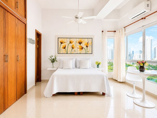 A bedroom with a bed, a ceiling fan, a window with a city view, and a wooden wardrobe.