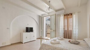 A bedroom with a bed, a TV on a cabinet, a chandelier, and a view of a balcony. - Tenuta Fioravante (Porto Cesareo)