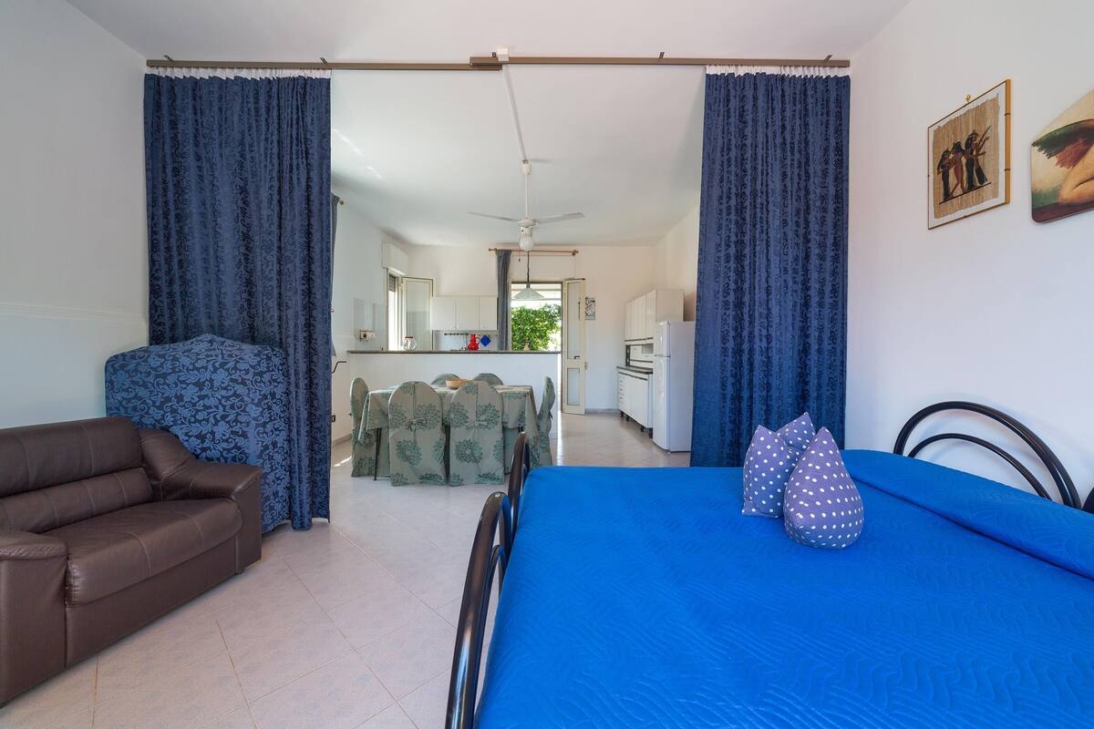 A room with a bed, a sofa, a dining table with chairs, and a kitchen area in the background.