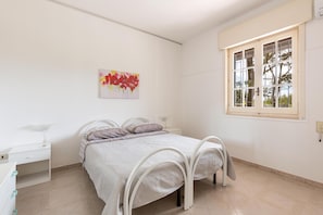 Une chambre à coucher comprenant un lit, une table de chevet, un bureau, une fenêtre avec des stores et une œuvre d’art accrochée au mur.