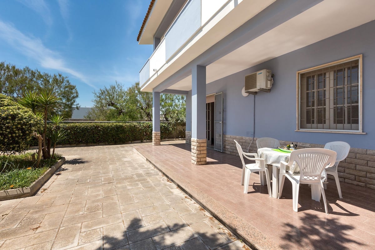 Eine überdachte Terrasse mit Tisch und Stühlen, ein überdachter Gang und ein Gebäude mit blauer Fassade.