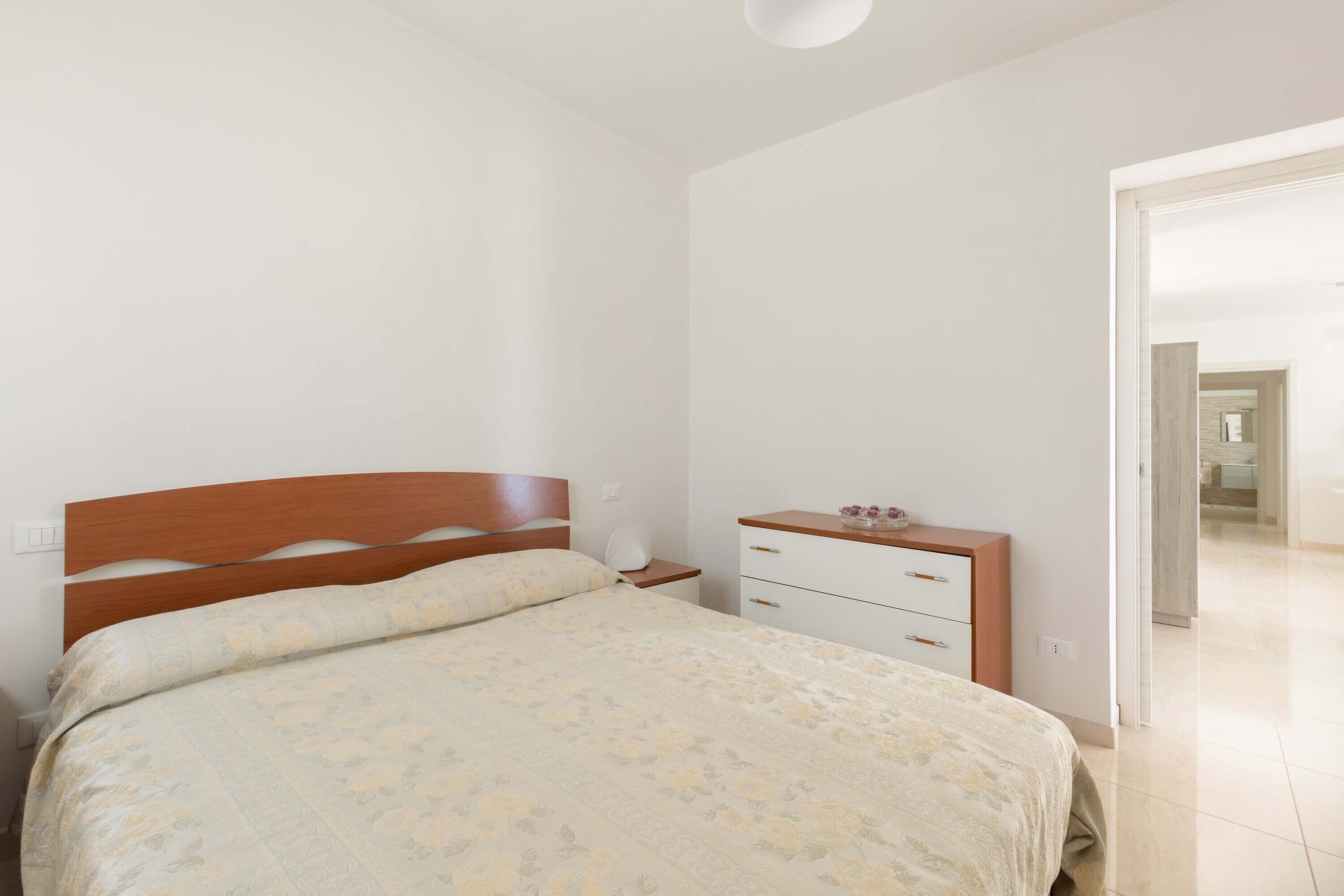 A bedroom with a wooden bed frame, a bedside table with a lamp, and a white dresser with drawers.