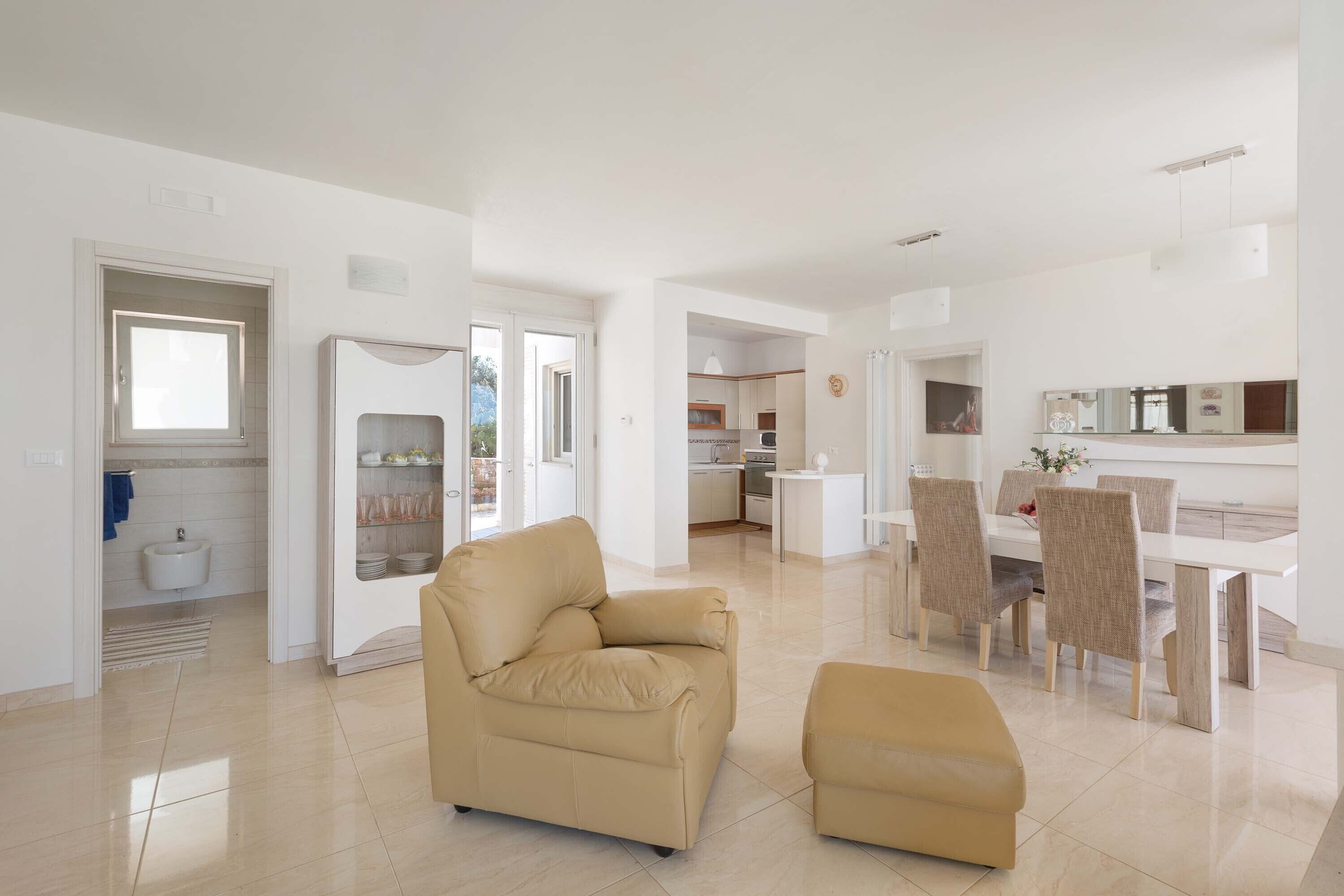 Un soggiorno moderno con un divano e poggiapiedi color beige, una zona pranzo con tavolo e sedie bianche, e una cucina sullo sfondo.
