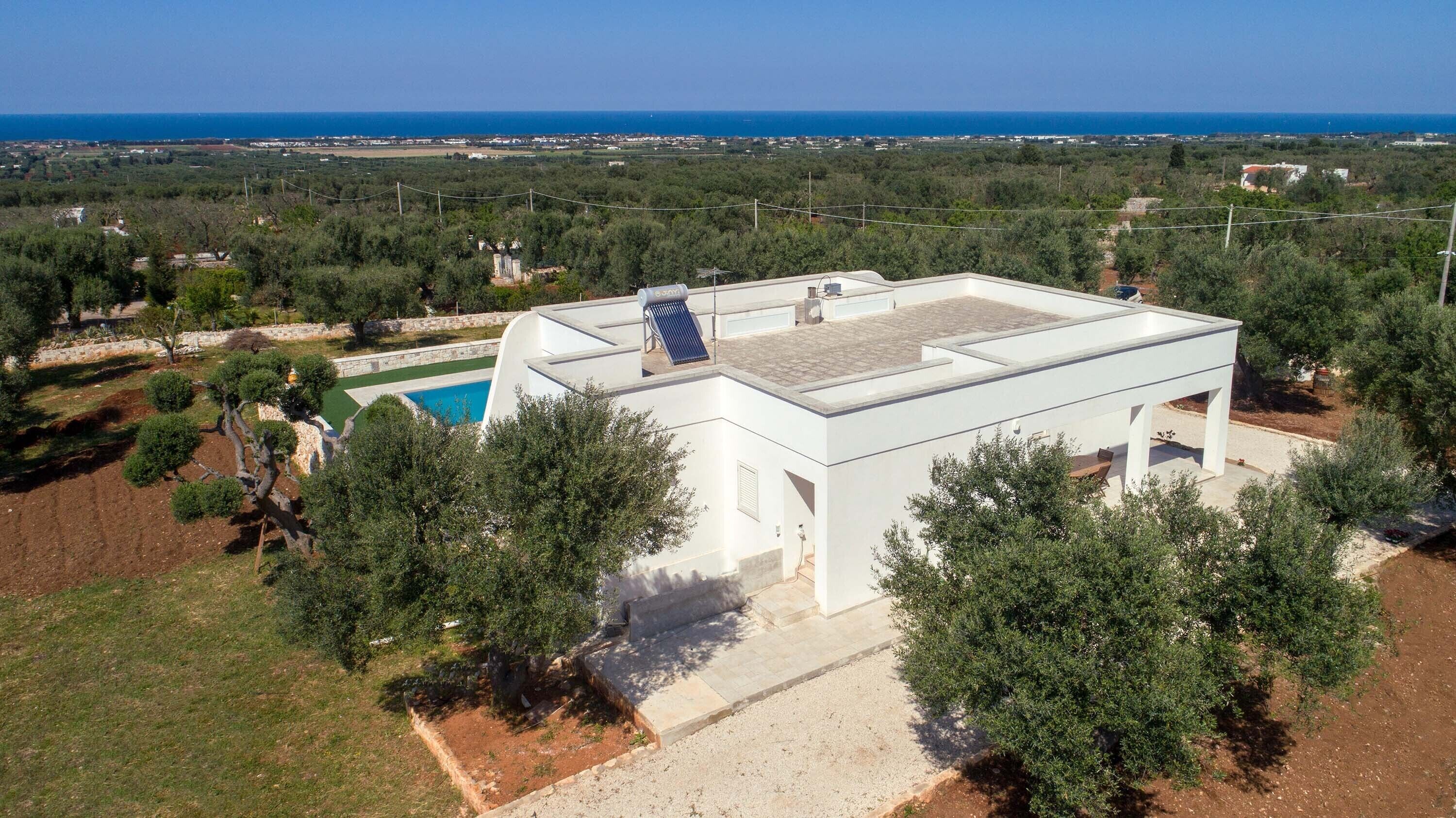 Una villa moderna blanca con piscina en la azotea y paneles solares, rodeada de vegetación y vistas al mar.