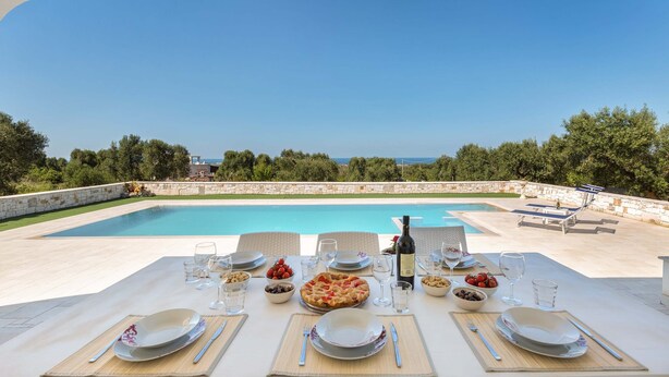 An outdoor dining area with a pool view, tables set with plates, glasses, and a bottle of wine. - Villa Specchia (Carovigno)