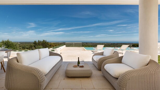 Un área de estar al aire libre techada con mobiliario de mimbre, una mesa con una botella y vasos, y vistas a una piscina y el paisaje.