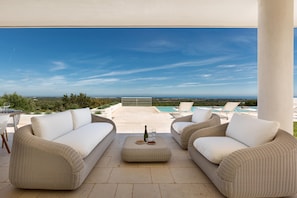 A covered outdoor seating area with wicker furniture, a table with a bottle and glasses, and a view of a pool and landscape. - Villa Orizzonte (Carovigno)