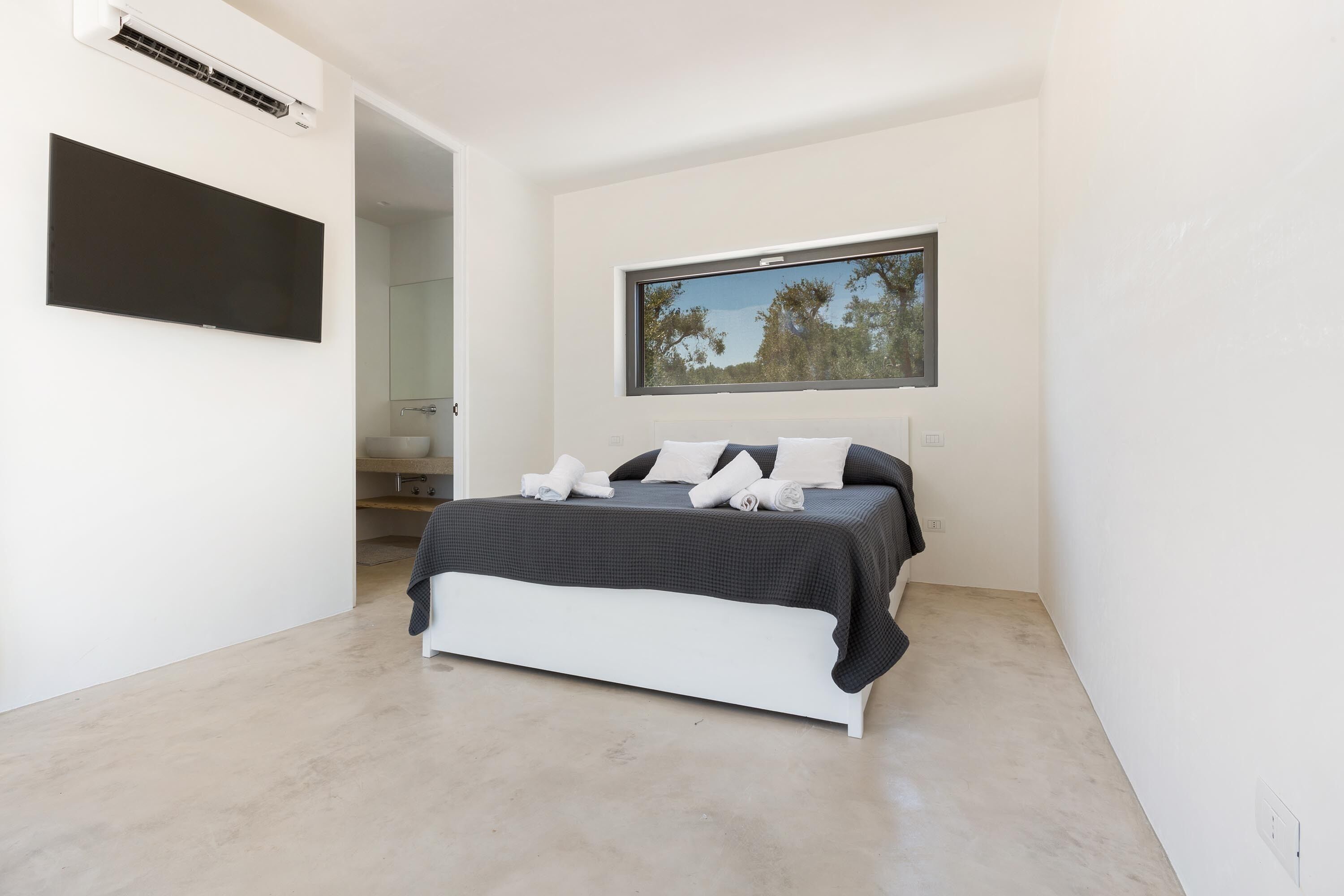 Un dormitorio moderno con una cama grande, un televisor de pantalla plana montado en la pared y una ventana con vistas a árboles.