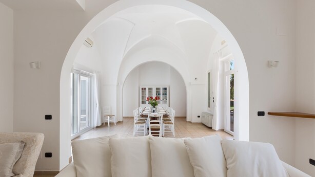 Een ruime woonkamer met een witte bank, een eettafel met stoelen en een grote, rondbogen doorgang naar een andere kamer.