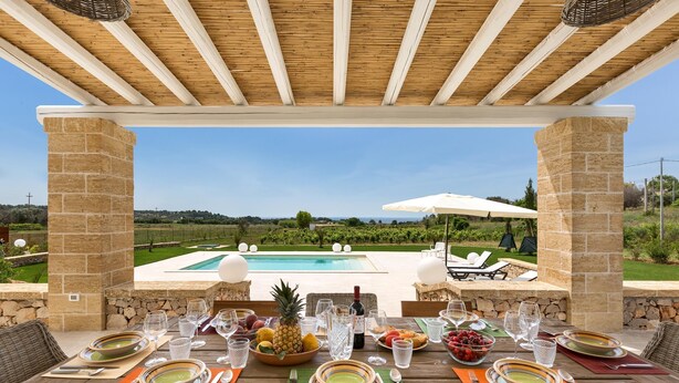 Un área de comedor al aire libre con vista a la piscina, pilares de piedra y una pérgola de madera.