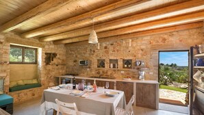A rustic stone interior with a dining area, stone walls, wooden beams, a stone bench, and a view through an open door. - Villa Elaion (Salve)