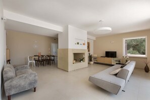 Uma sala de estar moderna com sofá, área de jantar com cadeiras e mesa, cozinha com televisão e uma janela ampla com vista para árvores.
