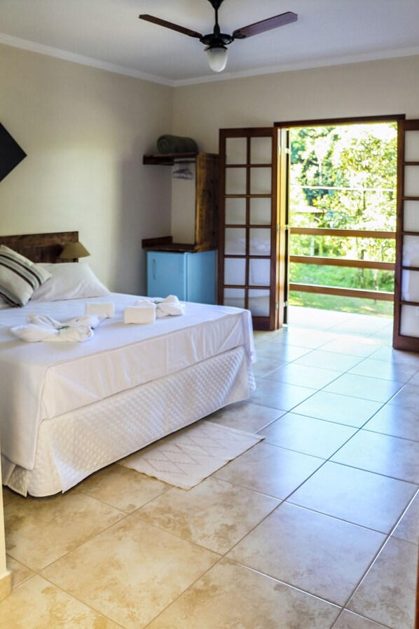 A hotel room with a bed, a ceiling fan, a window with a view of greenery, and a door leading to an outdoor area.