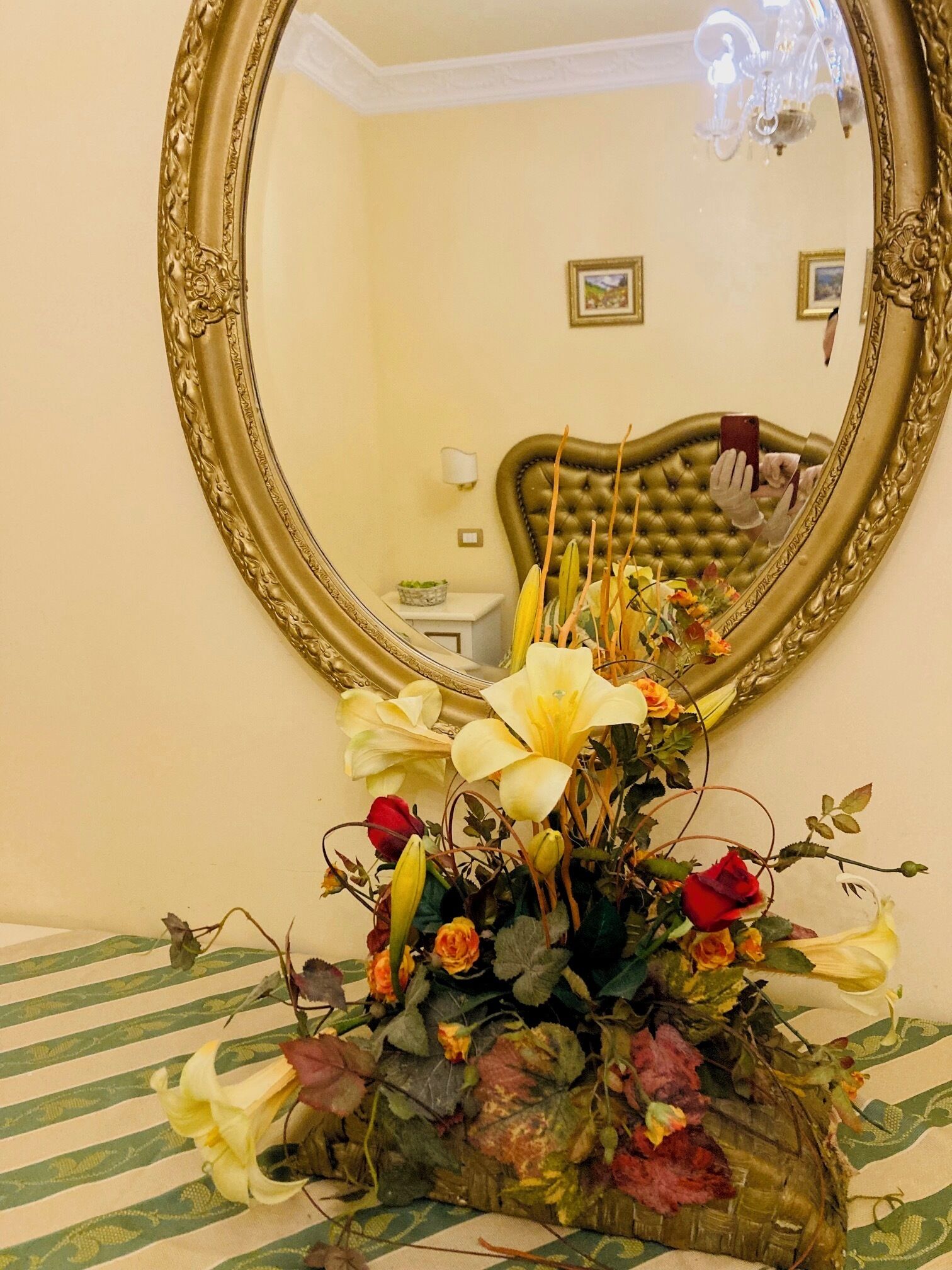 A mirror with an ornate gold frame, a floral arrangement in a vase, and a bed with striped bedding.
