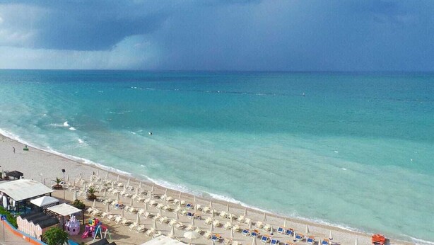 Une plage bordée de rangées de chaises longues et de parasols, un bâtiment doté d’une zone abritée, et une vue dégagée sur l’océan.