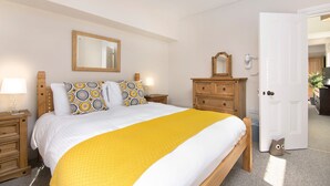 Ein ordentlich bezogenes Bett mit gelber Decke, Holz-Kopfstütze und gemusterten Kissen. Ein Nachttisch mit Lampe, eine Kommode und ein Spiegel an der Wand sind ebenfalls zu sehen.