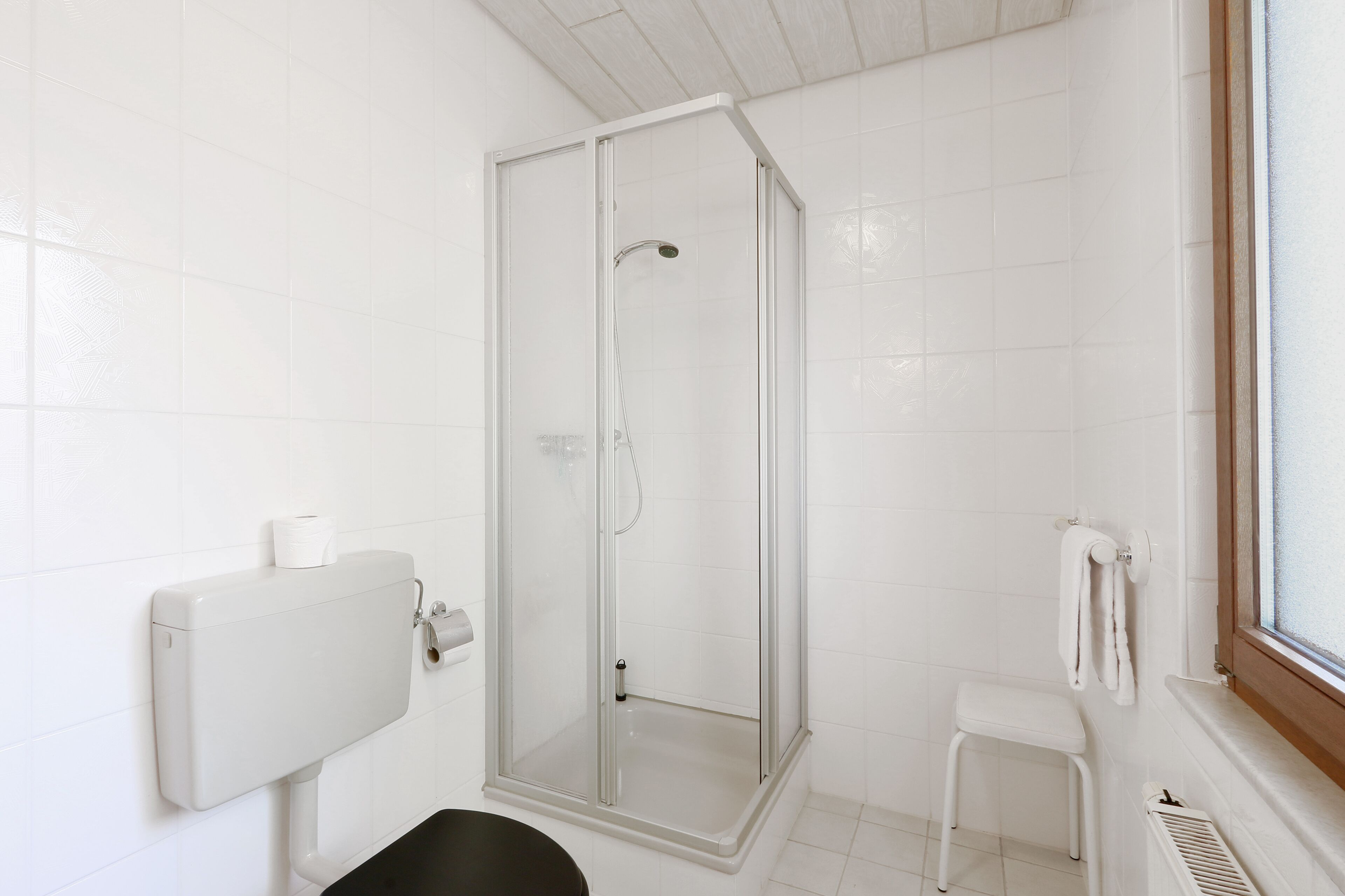Un baño con ducha acristalada, inodoro, un taburete pequeño y un perchero para toallas.