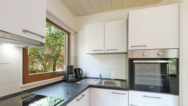 Cozinha moderna com armários brancos, bancada preta, forno embutido e janela com vista para árvores.