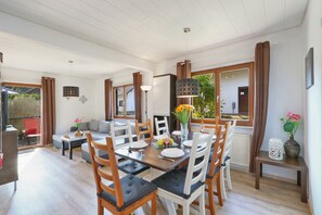 Zona pranzo con tavolo in legno e sedie bianche, divano e tavolo da pranzo apparecchiato con piatti, bicchieri e un vaso di fiori.