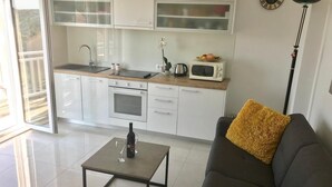 Eine moderne Küche mit weißen Schränken, einer Mikrowelle und einem Spülbecken. Ein Esstisch mit Flasche und Glas ist zu sehen. Im Vordergrund befindet sich ein Sofa mit einem gelben Kissen.