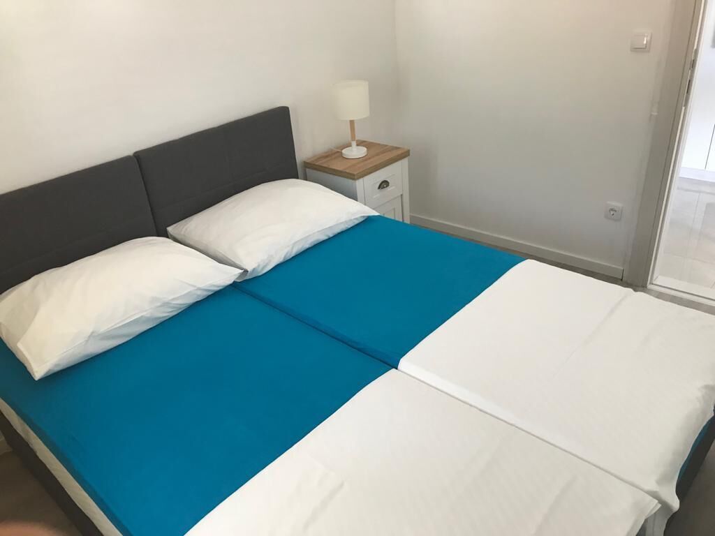 Una cama con colcha azul y blanca, una mesita de noche con lámpara, y una pared lisa.