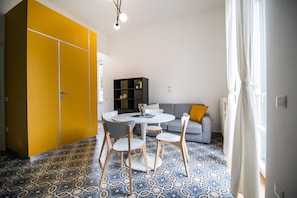 A room with a patterned floor, a round table with chairs, a sofa with cushions, and a bookshelf. - Salerno 1950 (Salerno)