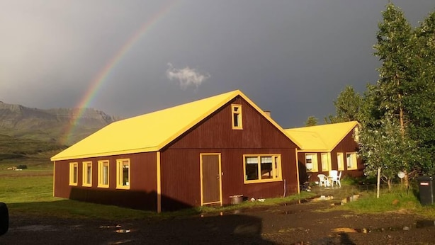 A wooden building with a yellow roof, a door, and windows, set in a rural landscape with a rainbow in the background. - Skarð (Fjardabyggd)